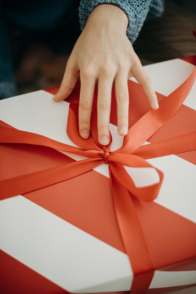 Dłoń trzymająca duży biały prezent owinięty czerwoną wstażką.