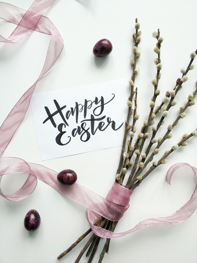 Napis Happy Easter na białej kartce, obok leży różowa wstążka i bazie.