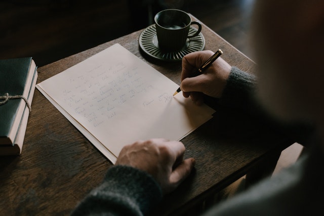 Osoba pisząca list, na stole stoi filiżanka oraz leży plik zeszytów.