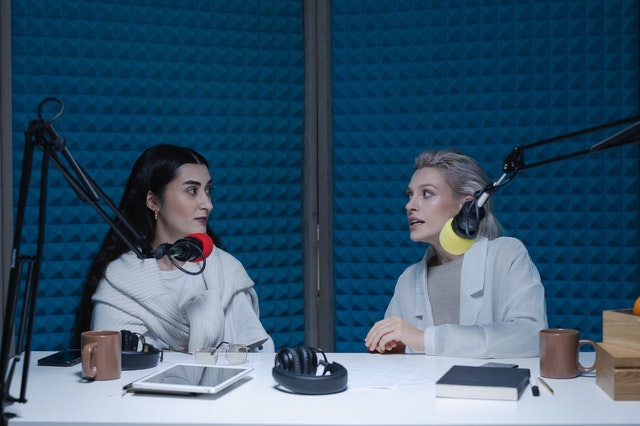 Na niebieskim w studiu radiowym siedzą dwie kobiety. Przed nimi znajdują się mikrofony.