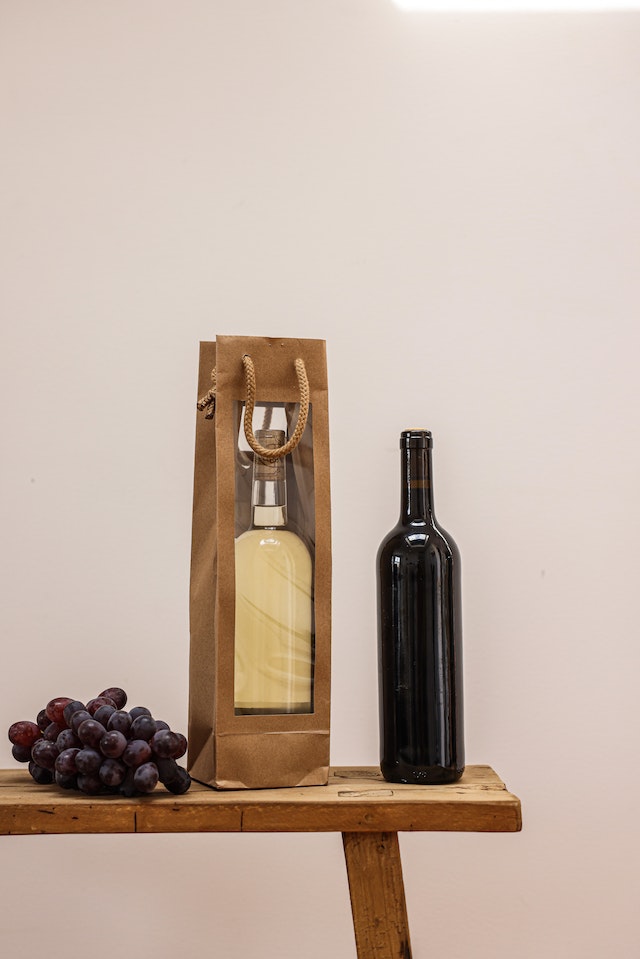 Butelka białego wina w ozdobnym opakowaniu, butelka ciemnego wina i ciemne winogrona na stole.