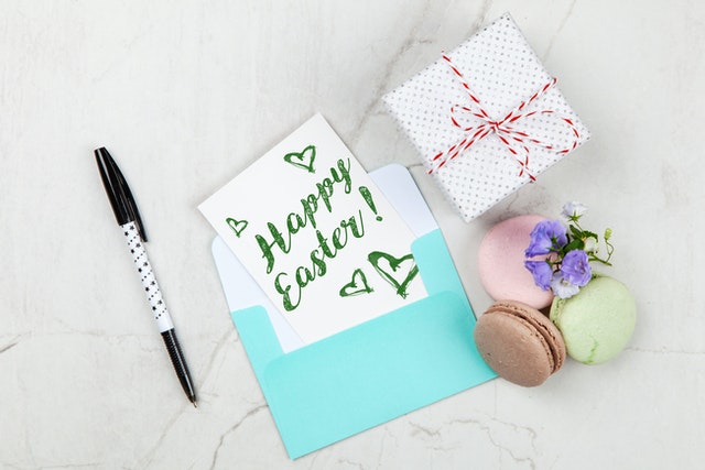 Napis Happy Easter na małej karteczce wychodzącej z miętowej koperty, mały prezent oraz trzy makaroniki i długopis.