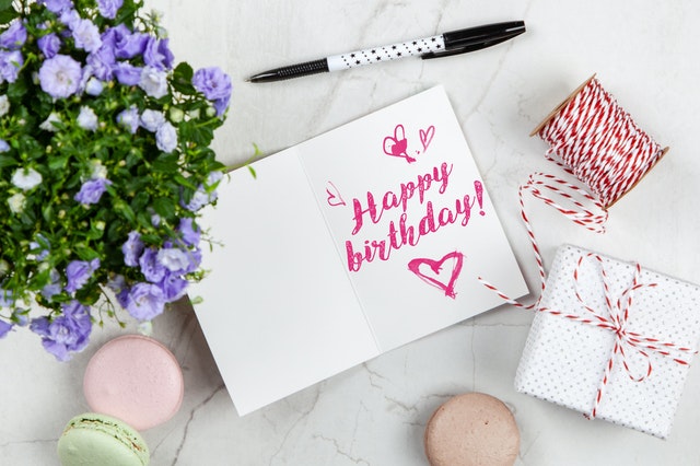 Karta z Happy birthday, obok leży zapakowany prezent, długopis i bukiet kwiatów.