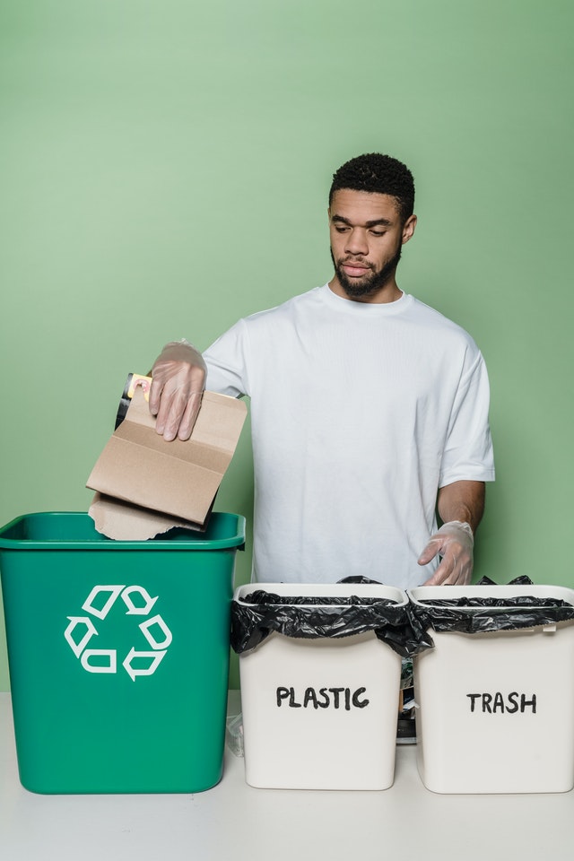 Mężczyzna segregujący śmieci. Przed nim stoją trzy kosze: plastic, trash i zielony z symbolem recyklingu.