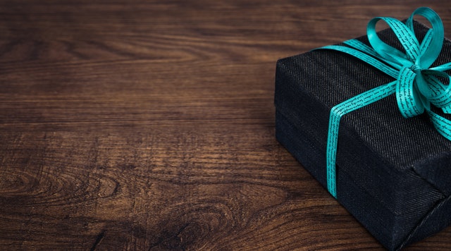 Ciemne pudełko prezentowe przewiązane błękitną wstążką na brązowym blacie.