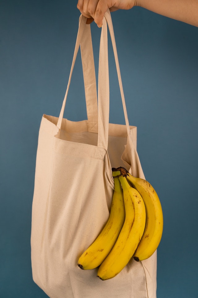 Ręka na niebieskim tle trzyma materiałową torbę. Z torby wystaje kiść bananów.